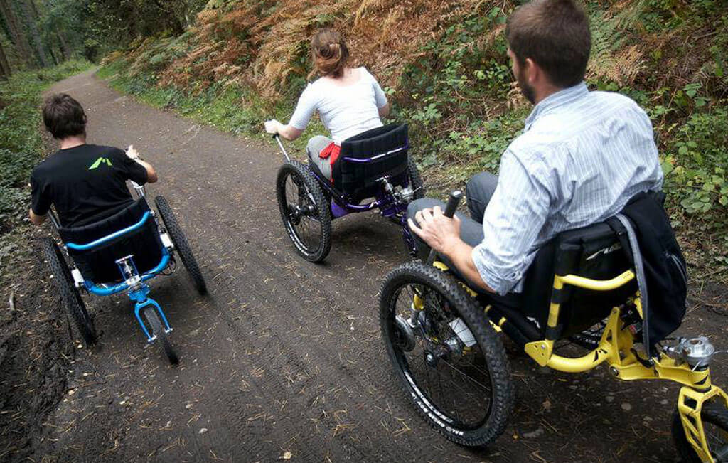 Tres personas utilizando sillas de ruedas todoterreno en un sendeiroflorestall.